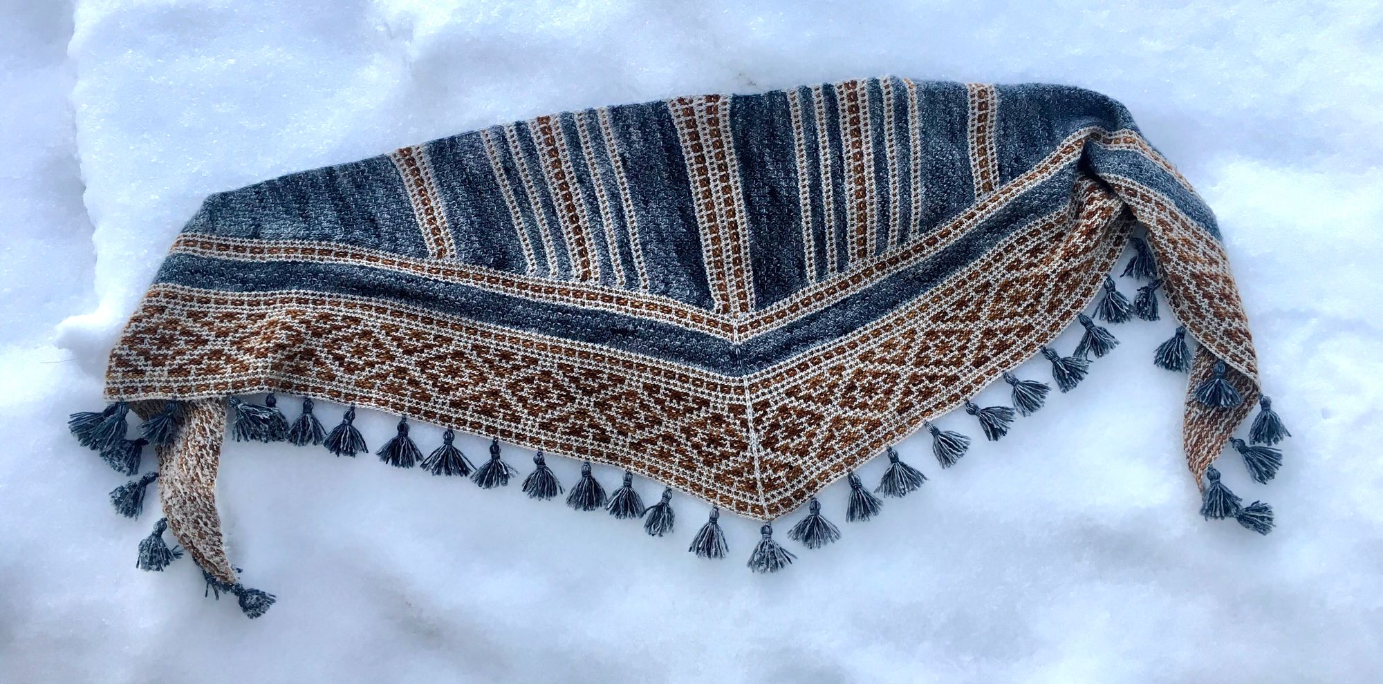 A tasseled shawl lying in the snow. The shawl is a mosaic pattern worked in a dusty blue yarn with gray and rust accent colors.